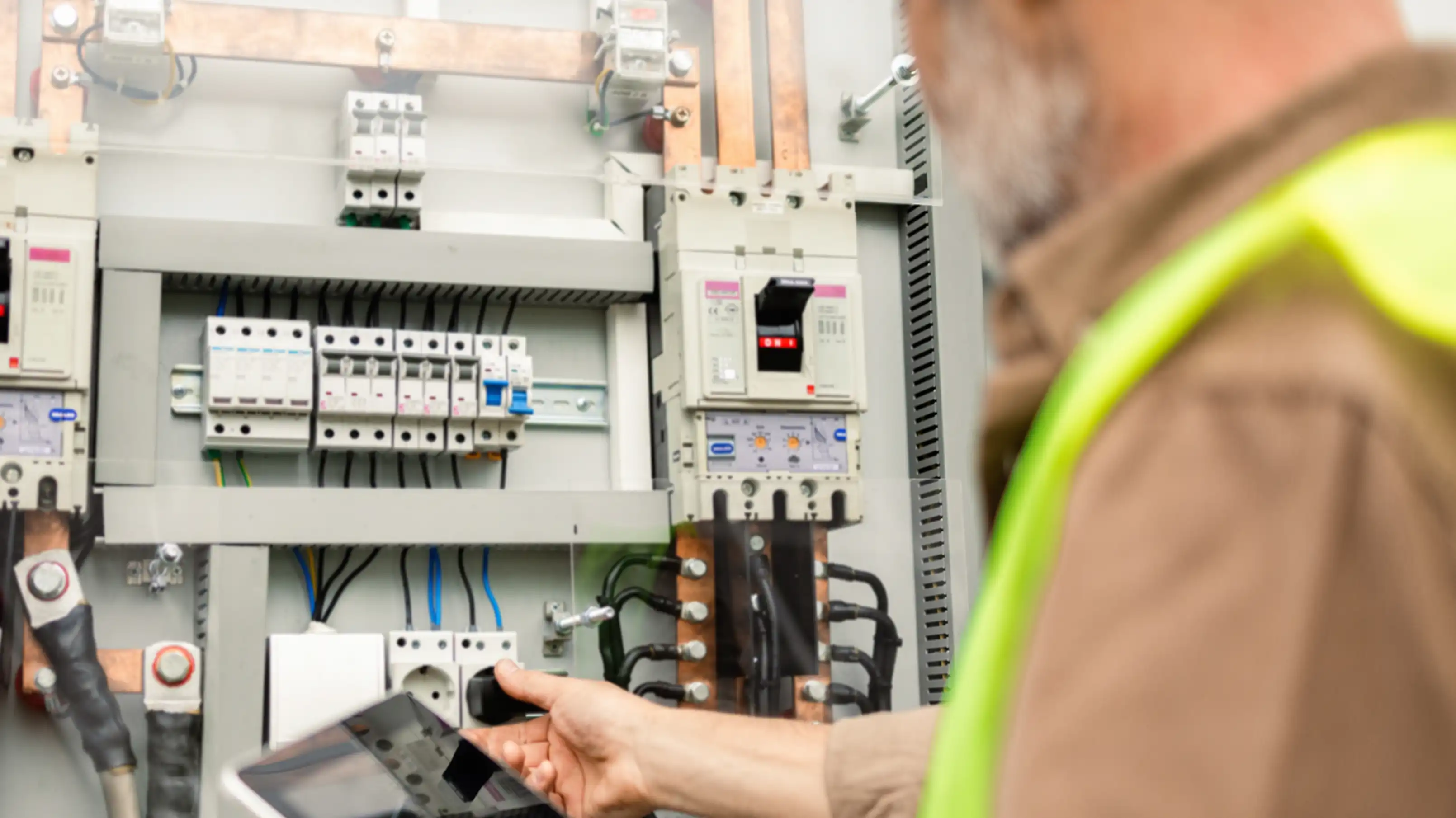 Fachkraft in Warnweste mit Tablet vor einem geöffneten Elektroverteilerschrank mit Sicherungsautomaten und Leistungsschalter.