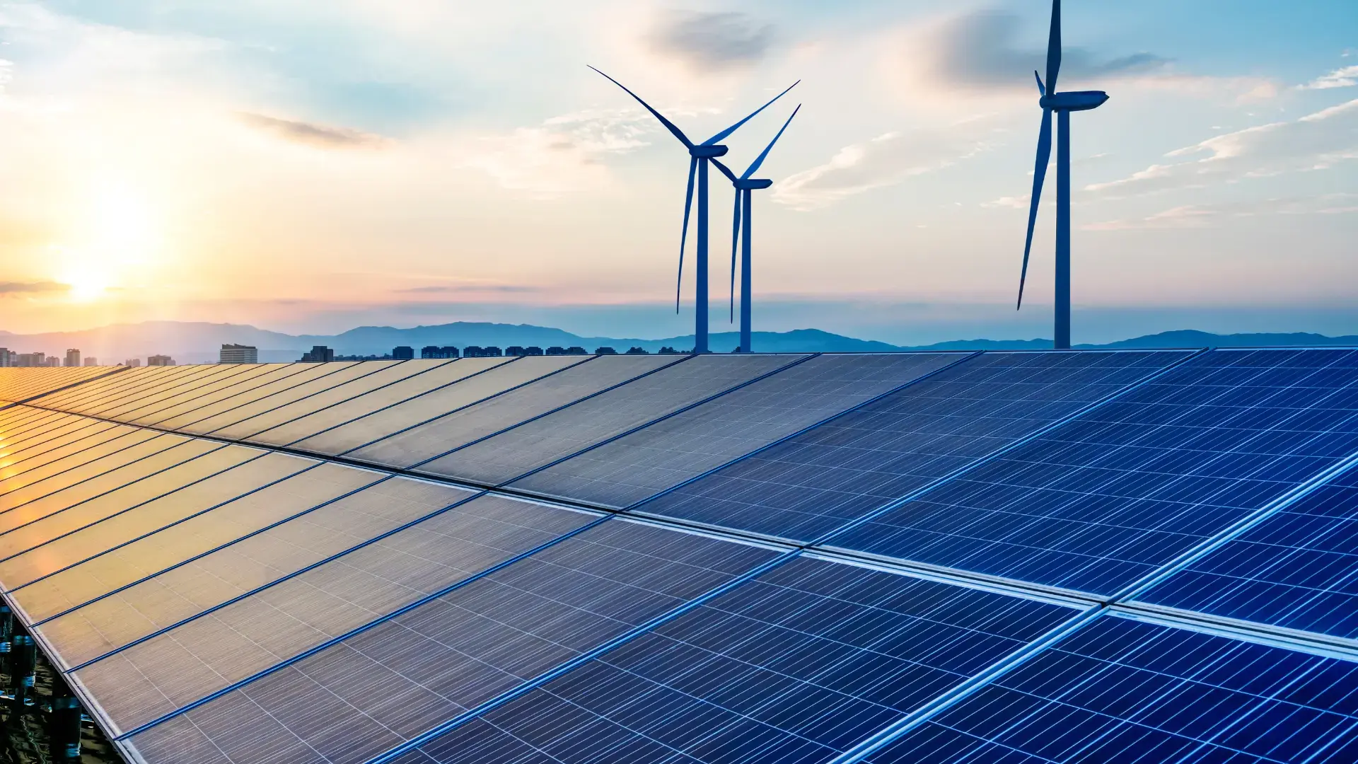 Photovoltaikmodule im Vordergrund, dahinter drei Windkraftanlagen vor einer Bergsilhouette im Abendlicht.